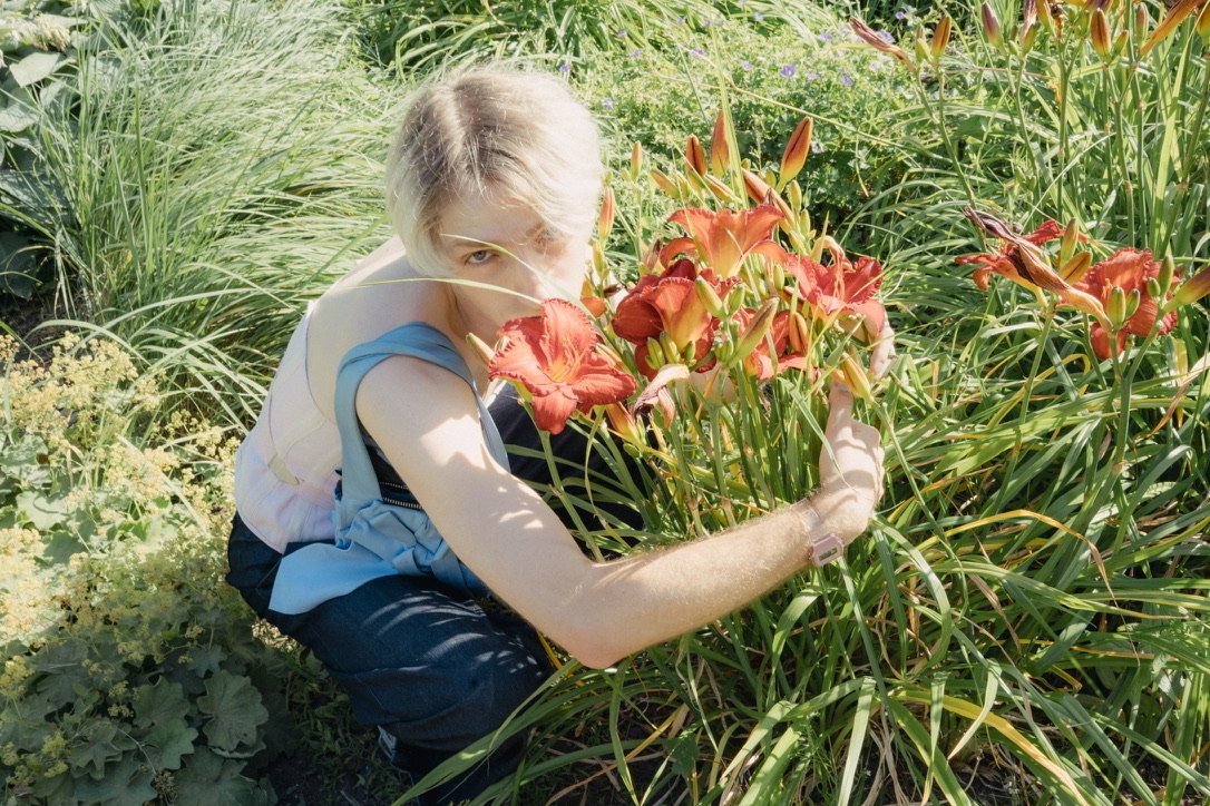 A person with light blond hair, wearing a sleeveless shirt, crouching among green plants and orange flowers, holding a bunch of orange flowers.