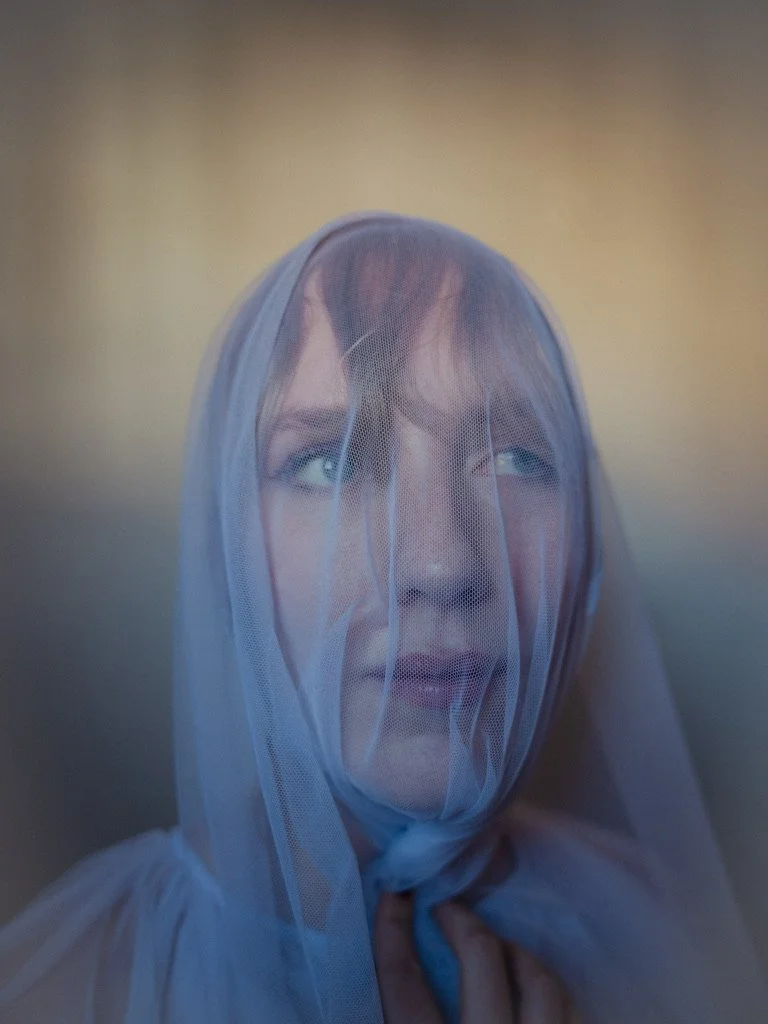 A woman with blue eyes and brown hair, wearing a white veil covering her face, looking upwards.