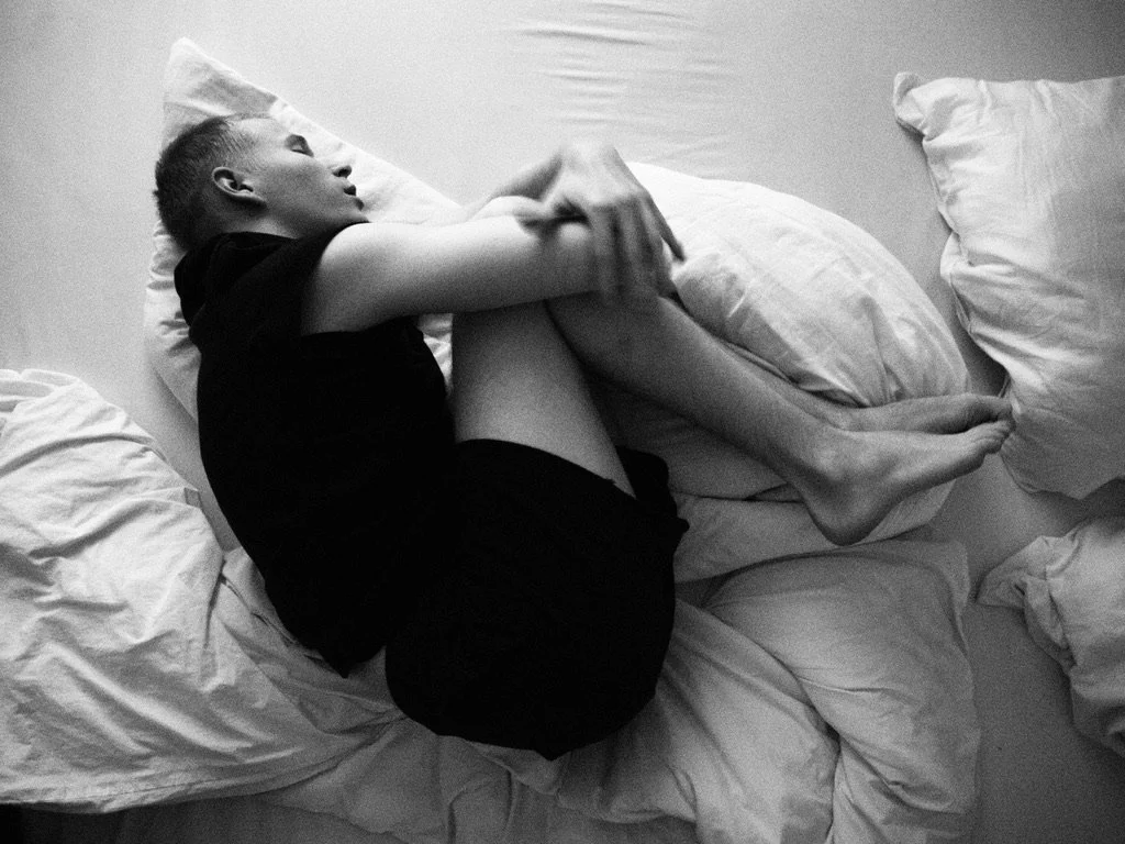 A person lying in bed on their side, holding their knees with their arms, with multiple pillows around them.