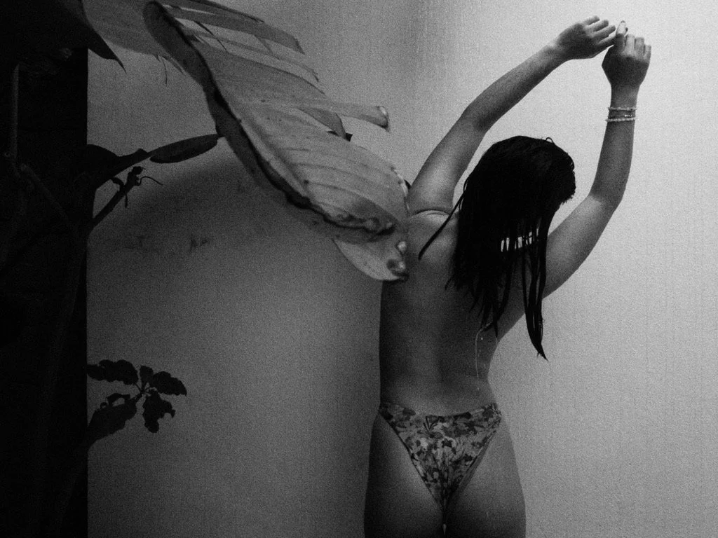 A woman with wet hair wearing patterned bikini bottoms, stretching her arms above her head, standing beside a large butterfly-shaped leaf and a plant with dark leaves.
