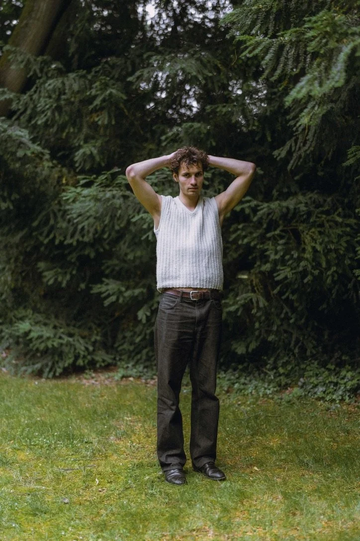A young man with curly hair wearing a sleeveless white top and black pants stands outdoors on a grassy area with trees and bushes in the background, holding his hands behind his head.