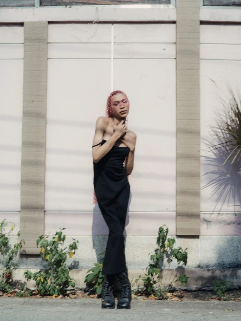 A woman with pink hair and dark lipstick standing against a wall with small plants at her feet, wearing a black dress and high-heeled boots.