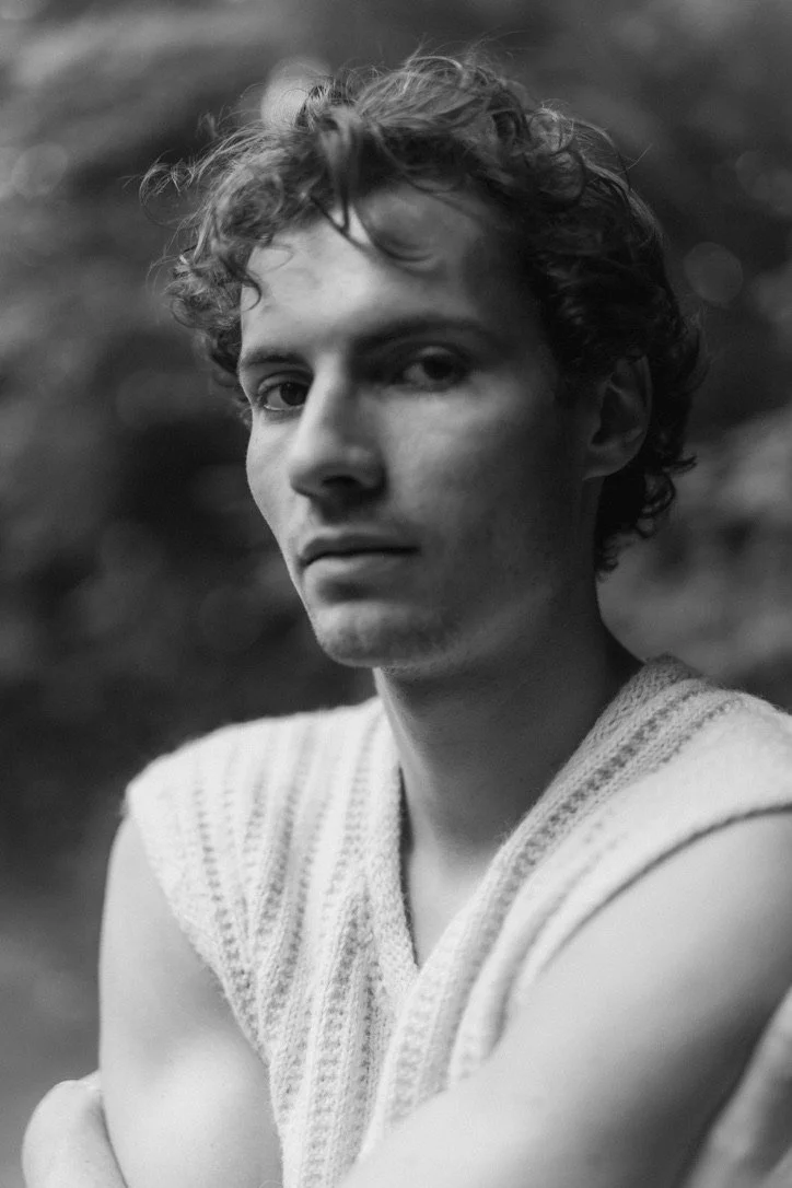 Black and white portrait of a young man with curly hair, wearing a sleeveless knit top, outdoors.