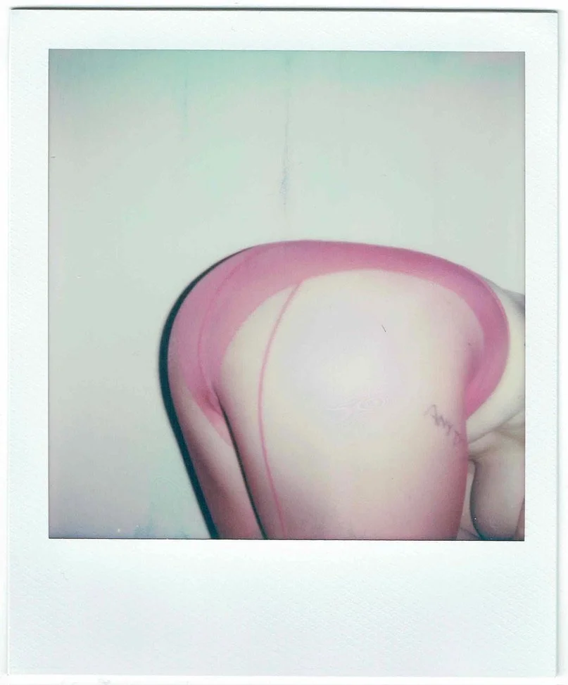 A person in pink underwear bending forward, showing their buttocks and thigh tattoo, against a plain white wall.