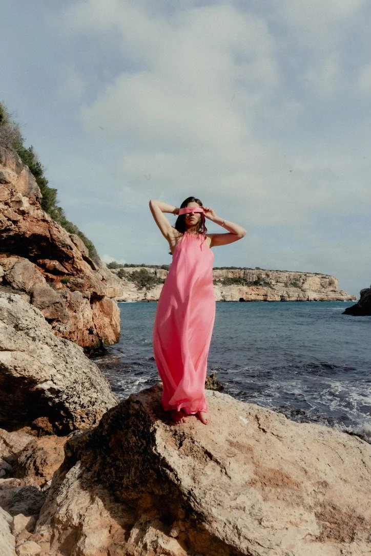 A woman in a long pink dress standing on a large rock by the water, shielding her eyes with her hand, at a rocky coastline on a partly cloudy day.