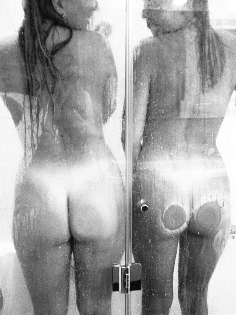 Two women taking a shower behind a foggy glass door, with one seen from the back and the other about to turn around.