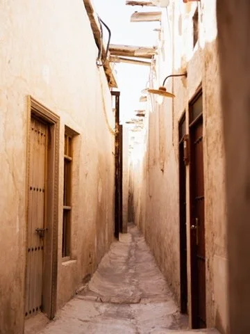 Narrow alleyway with old, beige stucco walls, small windows, and wooden doors on both sides, bathed in sunlight.
