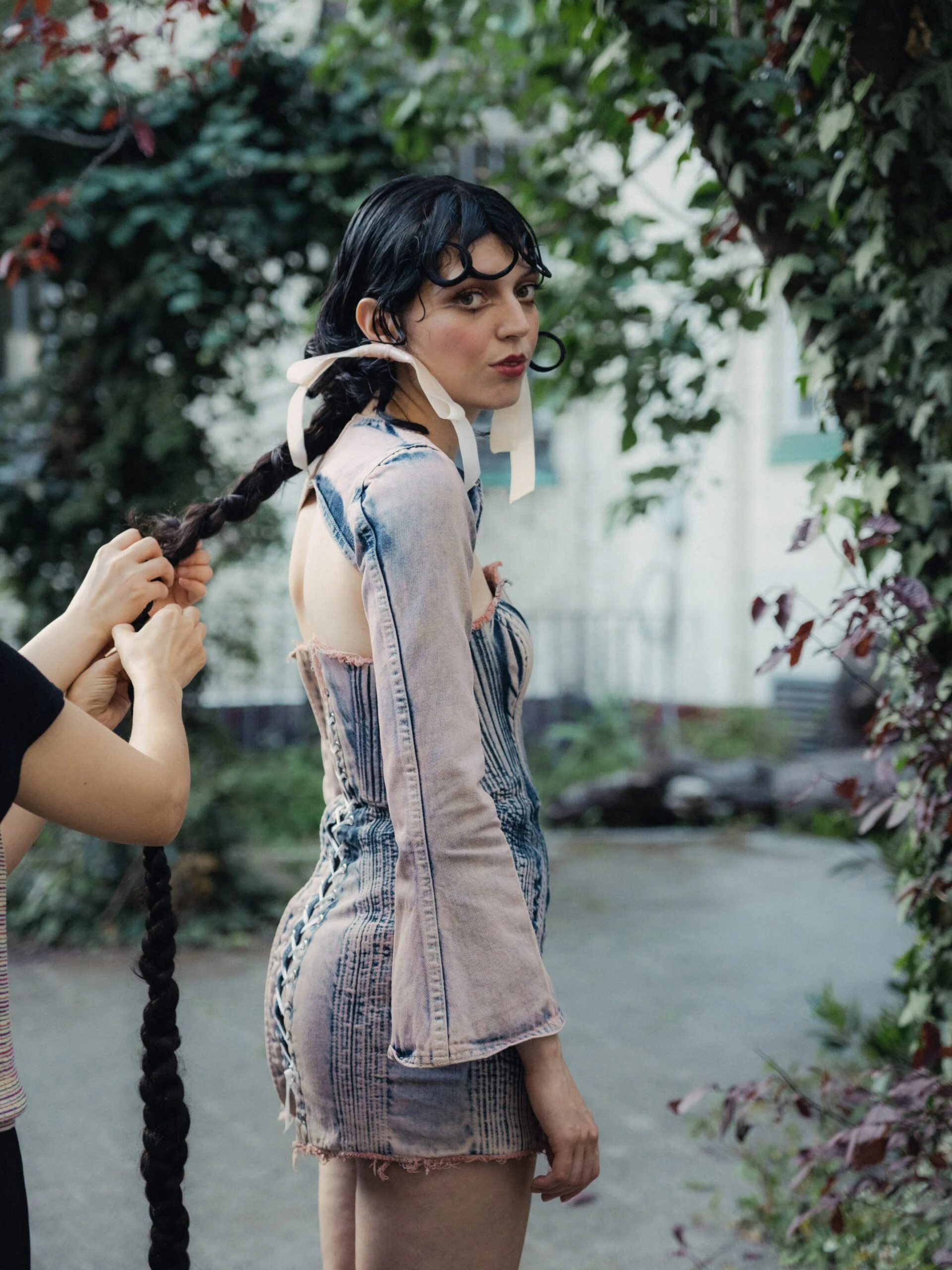 A young woman with dark hair styled with curl clips is standing outdoors, being styled into a braid by someone whose hands are visible. She is wearing a sleeveless, distressed denim jacket and shorts, surrounded by greenery and plants.
