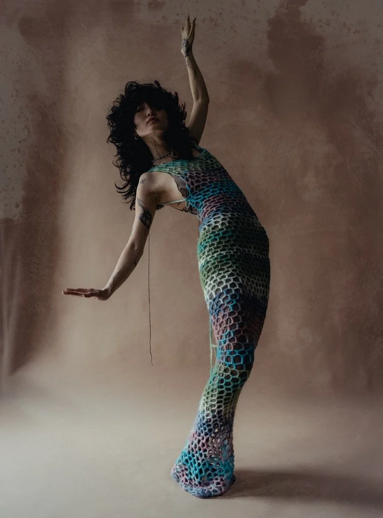 Woman with curly black hair wearing a colorful, netted dress poses with one arm raised and the other extended to the side against a textured brown background.