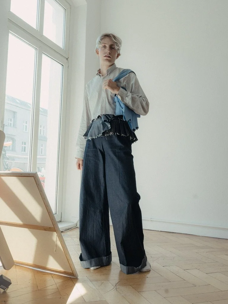 A person with short, light-colored hair standing inside a room near a large window, wearing a long sleeve striped shirt with frayed black denim shorts over wide-leg dark jeans, holding a blue garment over their shoulder.