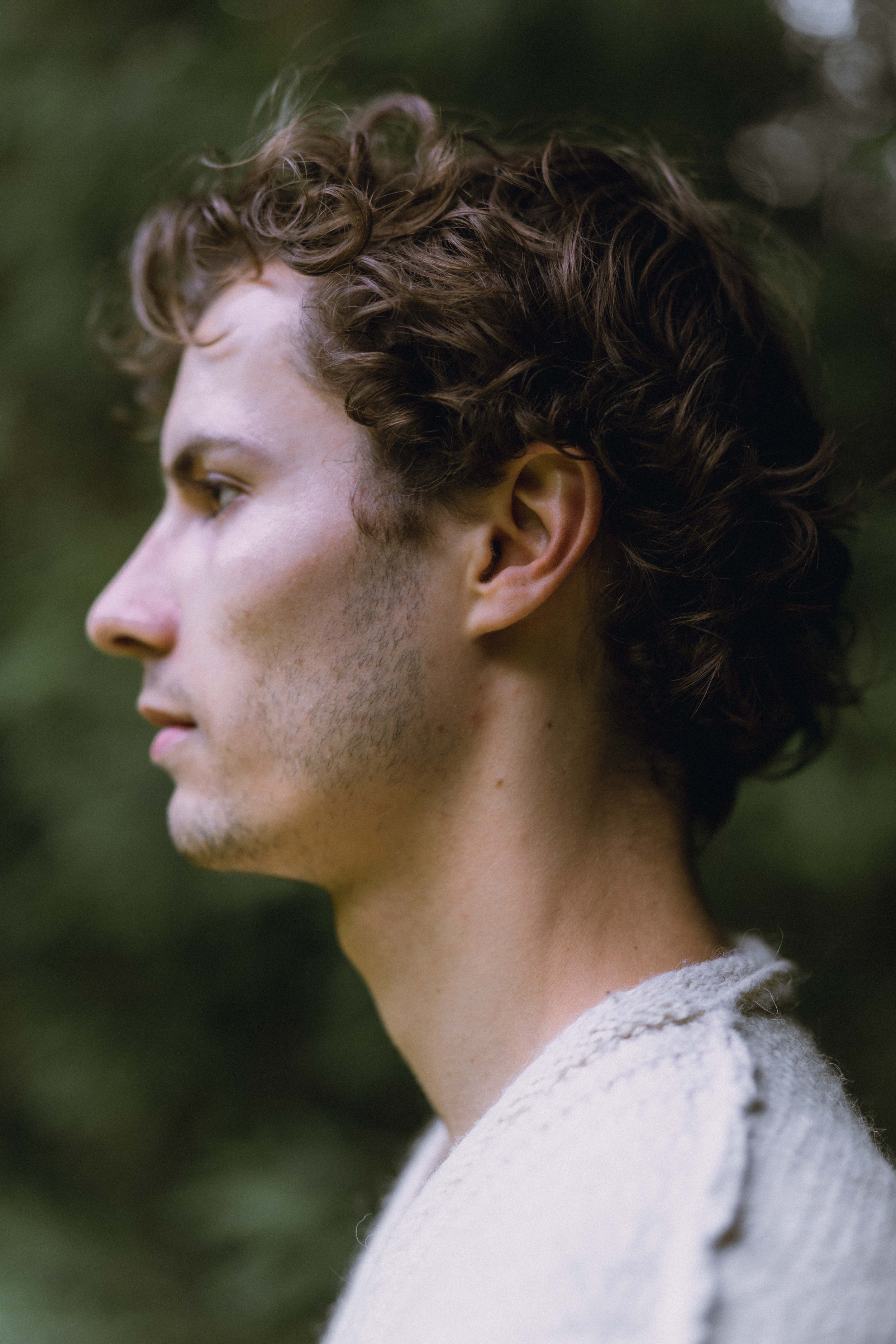 Side profile of a young man with curly brown hair, fair skin, closed eyes, and a neutral expression, wearing a light-colored sweater, outdoors with blurred green foliage in the background.