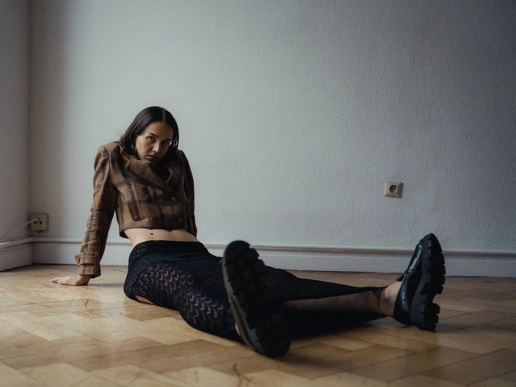 A woman is sitting on a hardwood floor against a plain white wall, wearing a cropped brown plaid jacket, black patterned leggings, and hiking boots, with a serious expression and looking at the camera.