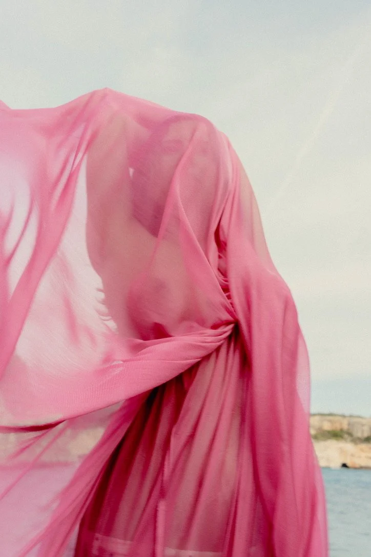 Close-up of a person dressed in a flowing, semi-transparent pink fabric, standing outdoors near water with a rocky landscape in the background.