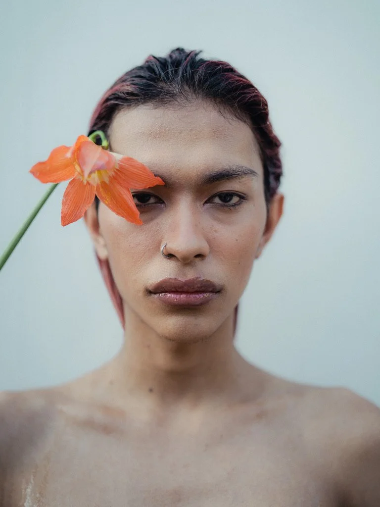 A young person with short dark hair, wearing a nose ring, holds an orange flower next to their face, looking directly at the camera against a plain neutral background.