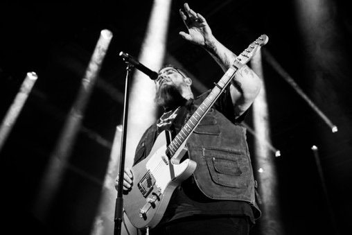 Musician with a guitar performing on stage in black and white, with stage lights in the background.