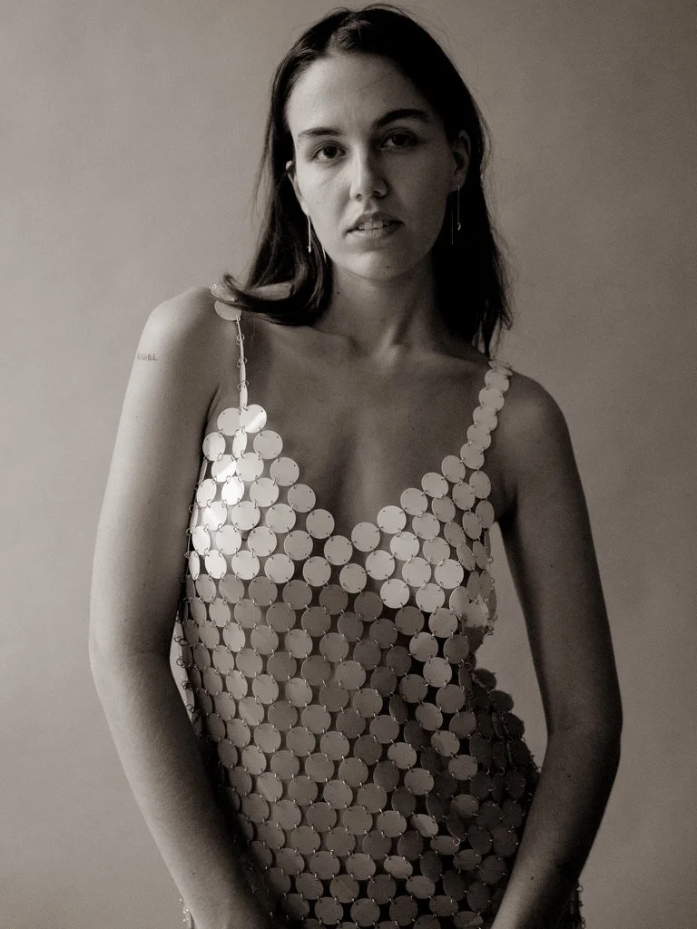 A woman with shoulder-length dark hair wearing a sleeveless dress made of large reflective circular discs standing against a plain light-colored background.