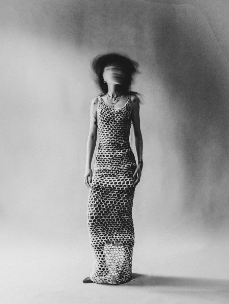 A black-and-white photo of a woman wearing a long, patterned dress. Her head is blurred, creating a sense of motion.