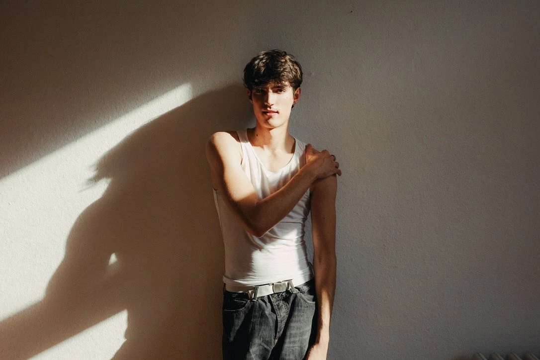 Young man with dark hair standing against a beige wall, partially illuminated by sunlight, wearing a white sleeveless shirt and dark pants.