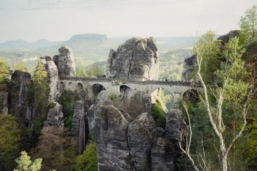 A stone bridge with multiple arches spanning across large rocks in a forested landscape.