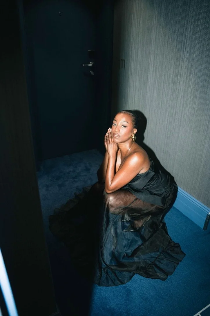 A woman in a black satin gown kneels on a blue carpeted floor with her hands cupping her face, in a dimly lit room with gray wallpaper and a dark door in the background.