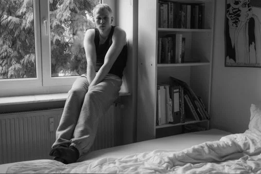 Person sitting on windowsill in a bedroom, looking at the camera, with a bed and bookshelves in the background.