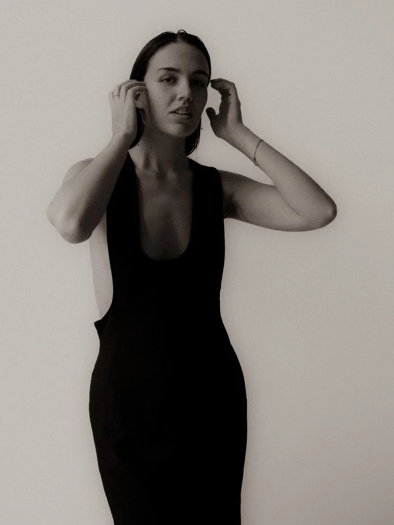 A woman with dark hair wearing a sleeveless black dress, standing against a plain white wall, touching her ears with both hands.