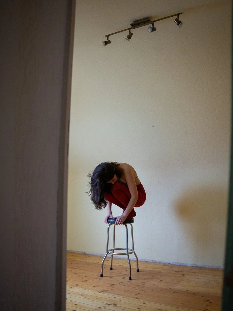 A woman crouches on a stool, holding onto the top, in a room with wooden flooring and cream-colored walls, with track lighting on the ceiling.