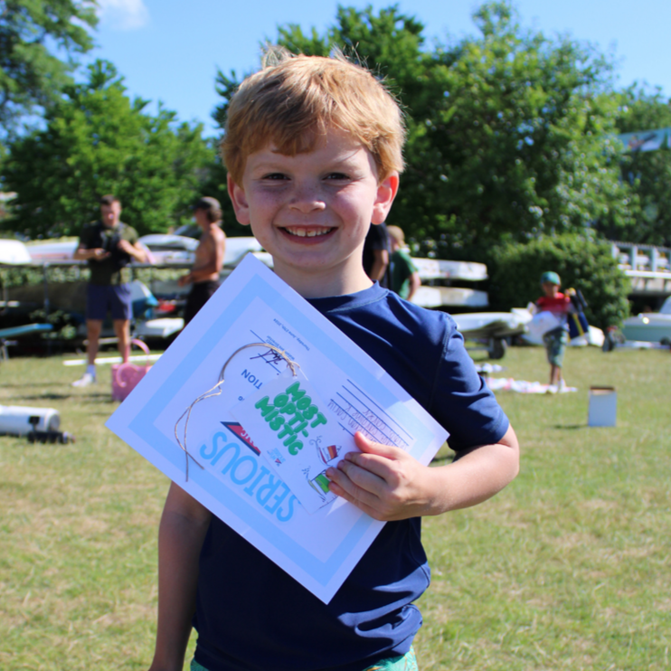 A young boy with red hair, smiling, holding a certificate at an outdoor event on a sunny day, with other children and adults in the background. He wins an award at Start Opti Sailing.