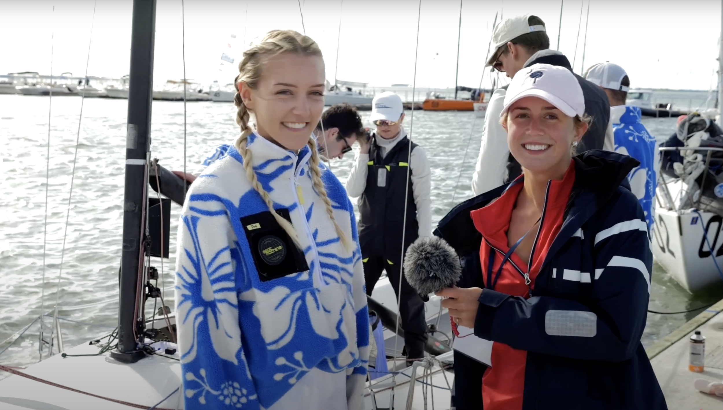 Two women, Marina Lamphier, a sailor, and Carly Starck, an interviewer, are smiling near sailboats at a marina. One of them is holding a microphone, while a group of people is preparing boats in the background.