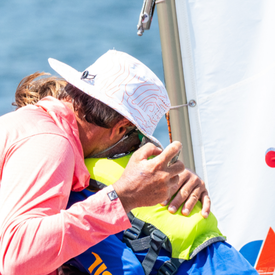 Dad, wearing a white hat and pink jacket, leans over and congratulates his child, who is wearing a life jacket and sitting on a boat, on winning his opti championship race in Minnetonka, Minnesota.