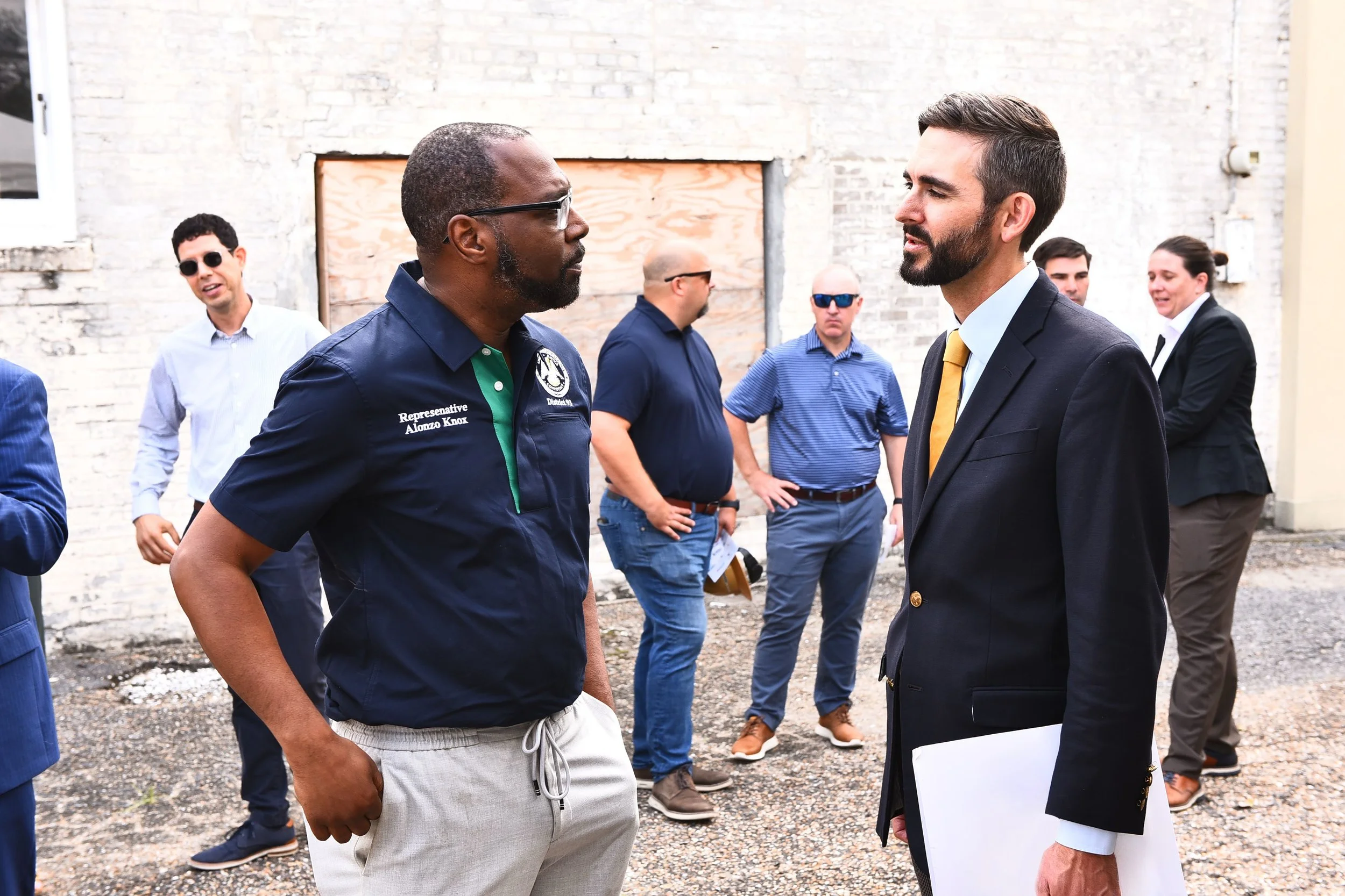 Two men in conversation outdoors, one wearing a navy shirt with patches and the other in a black suit with a yellow tie, with a group of people in the background near a brick wall.
