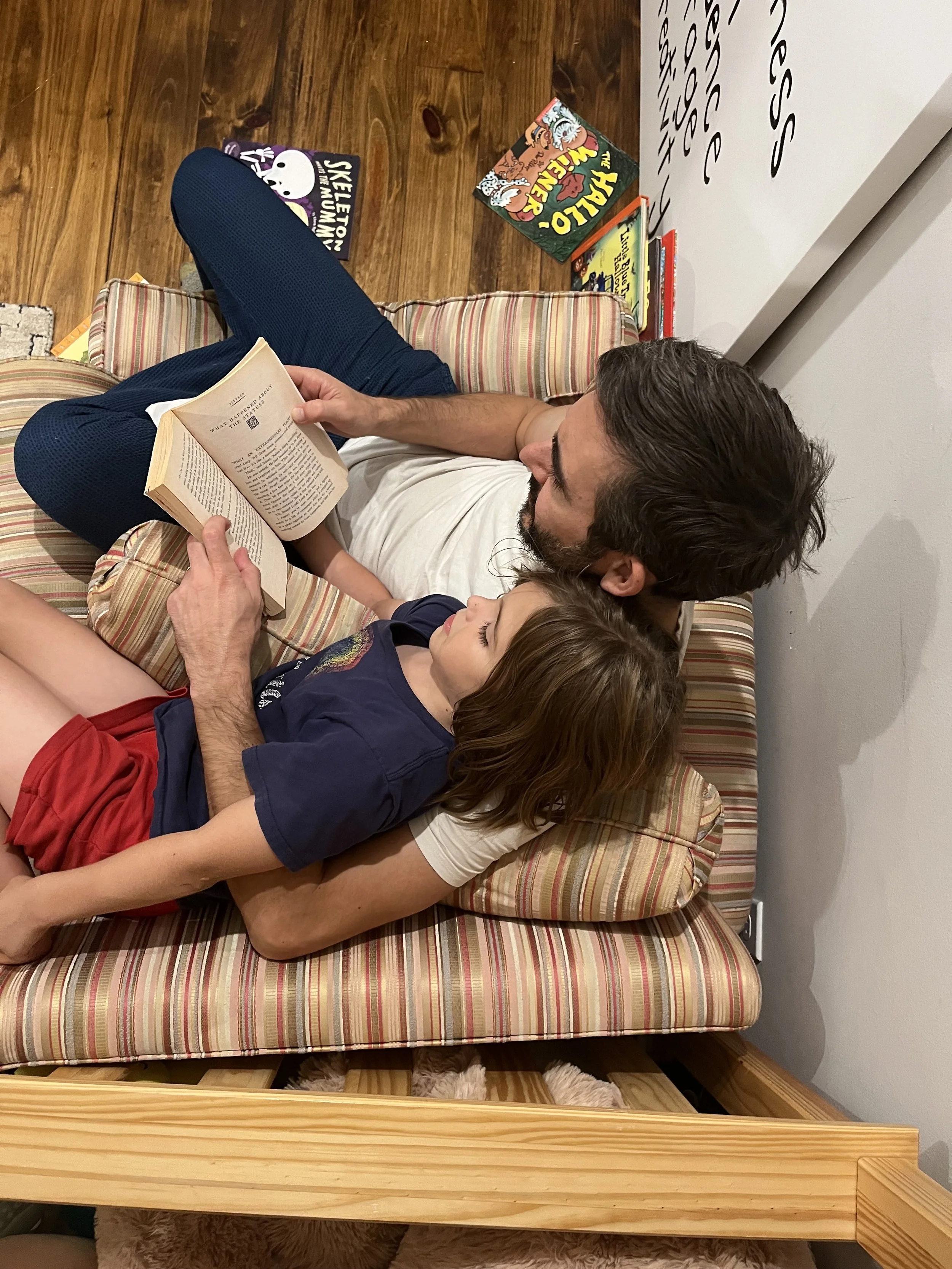 A man and a young girl lie on a striped cushioned bench, reading a book together, with books on the floor nearby.