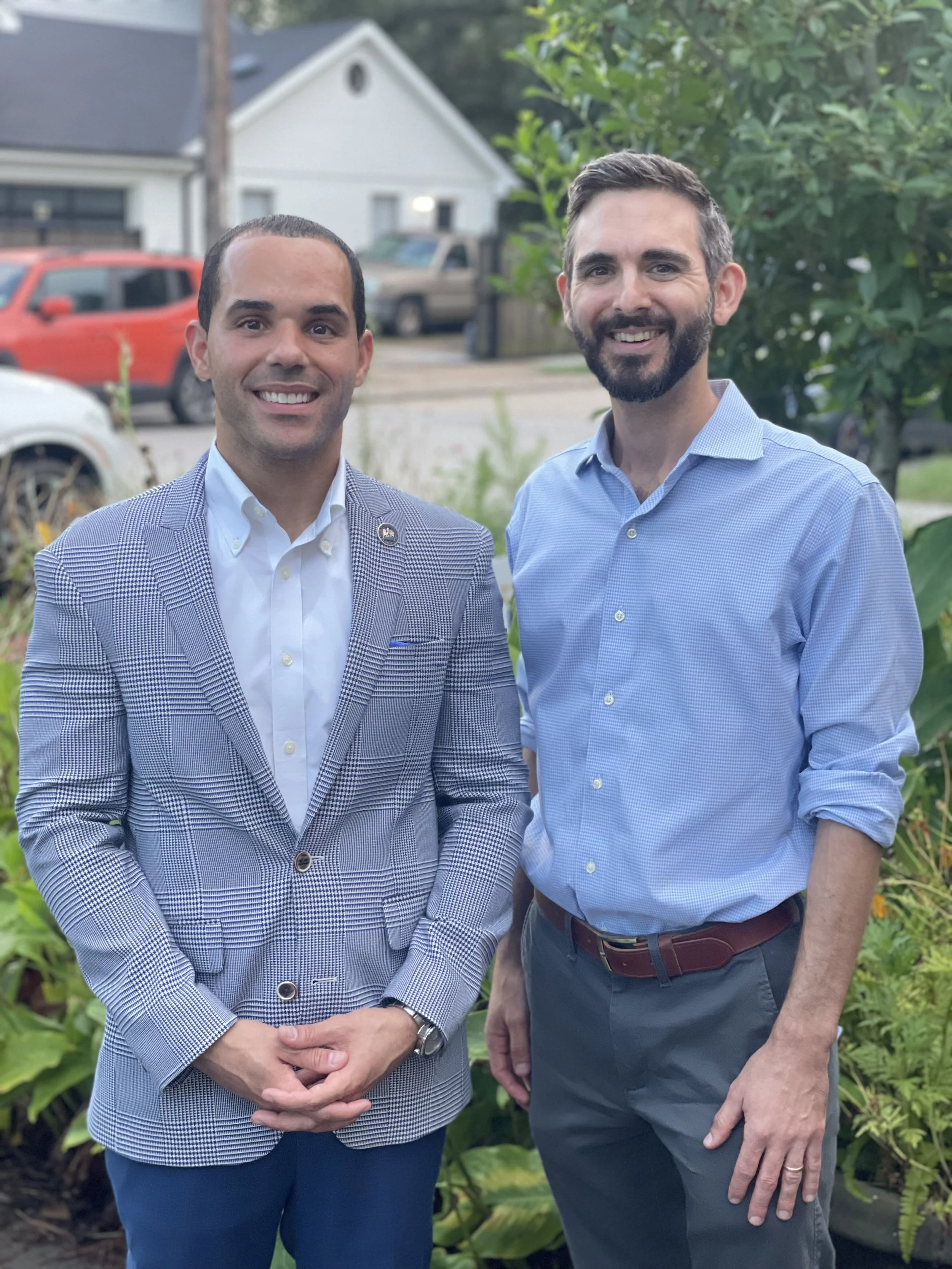 Two men standing outdoors, smiling, with trees and houses in the background.
