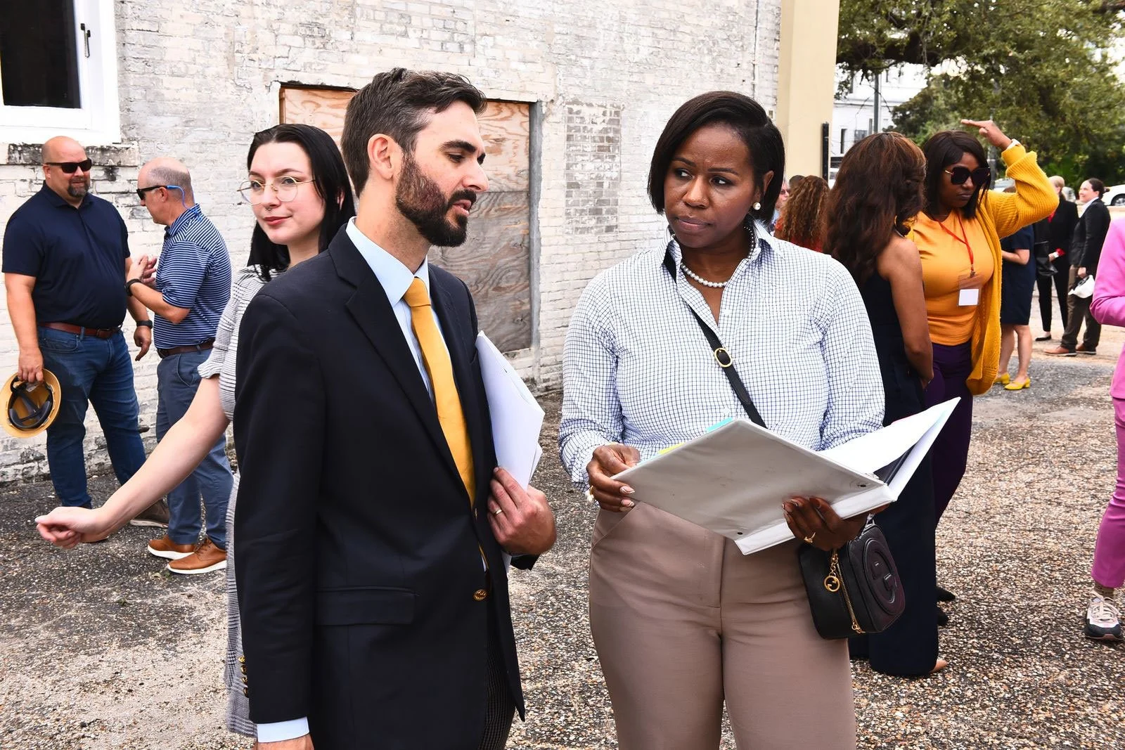 Two professionally dressed women and a man engaging in conversation outdoors at a gathering, with other attendees in the background.