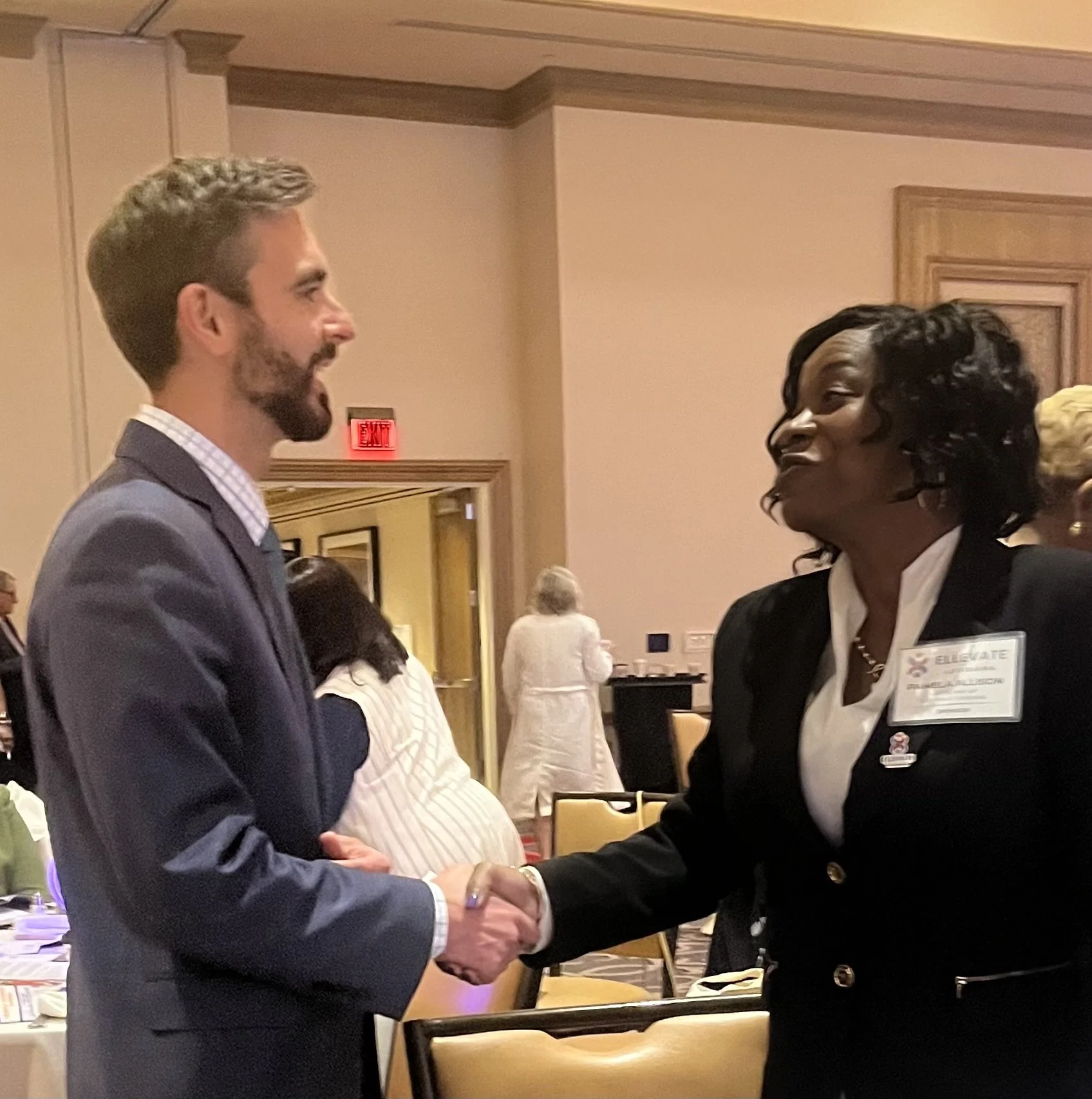 A man and a woman shaking hands at a formal event inside a room with beige walls and wooden trim. The man has light skin, a beard, and is wearing a grey suit with a checkered shirt. The woman has dark skin, curly hair, and is dressed in a black blaze