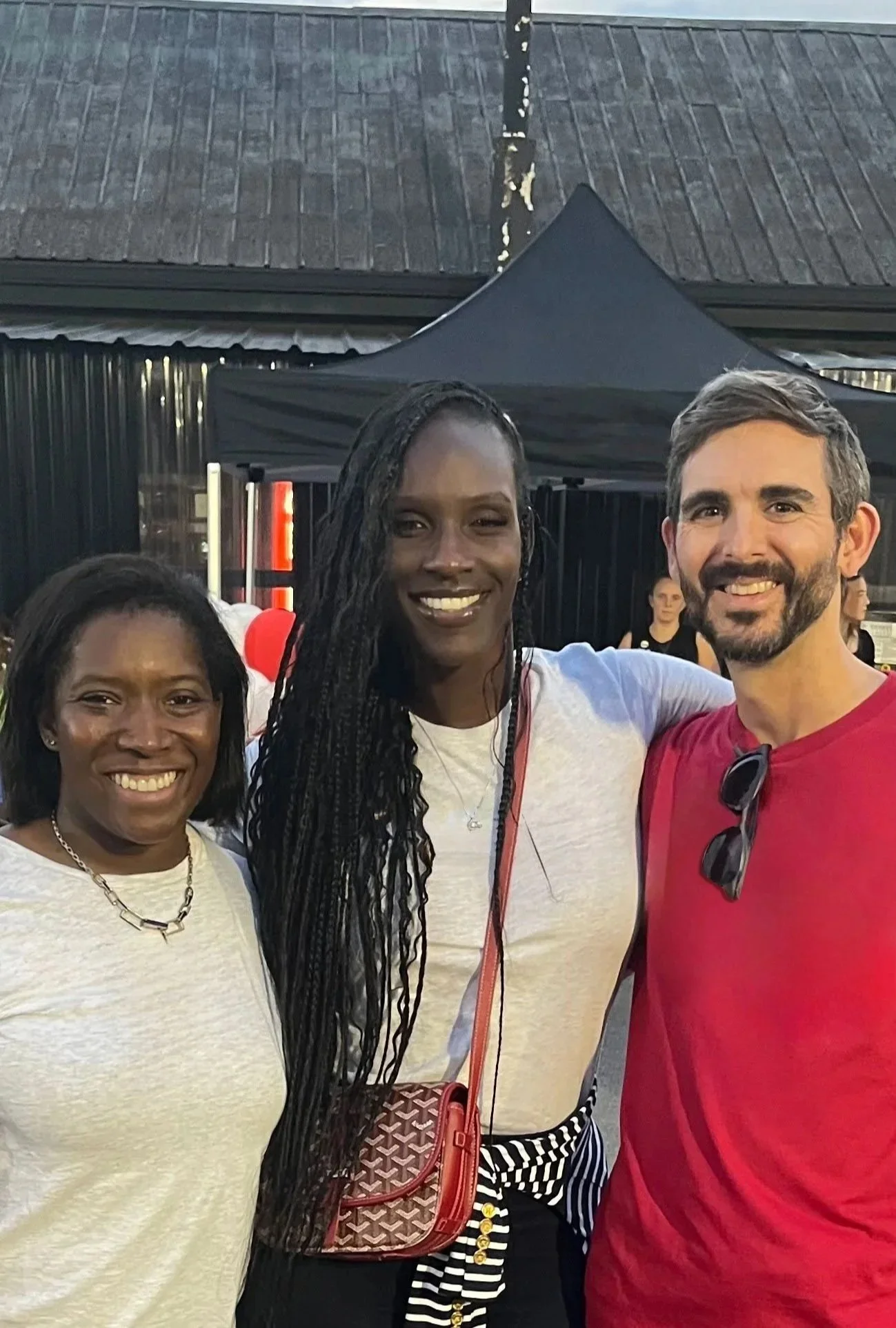 Three people smiling and posing outdoors at a social event, with a black canopy and a building with a sloped roof in the background.