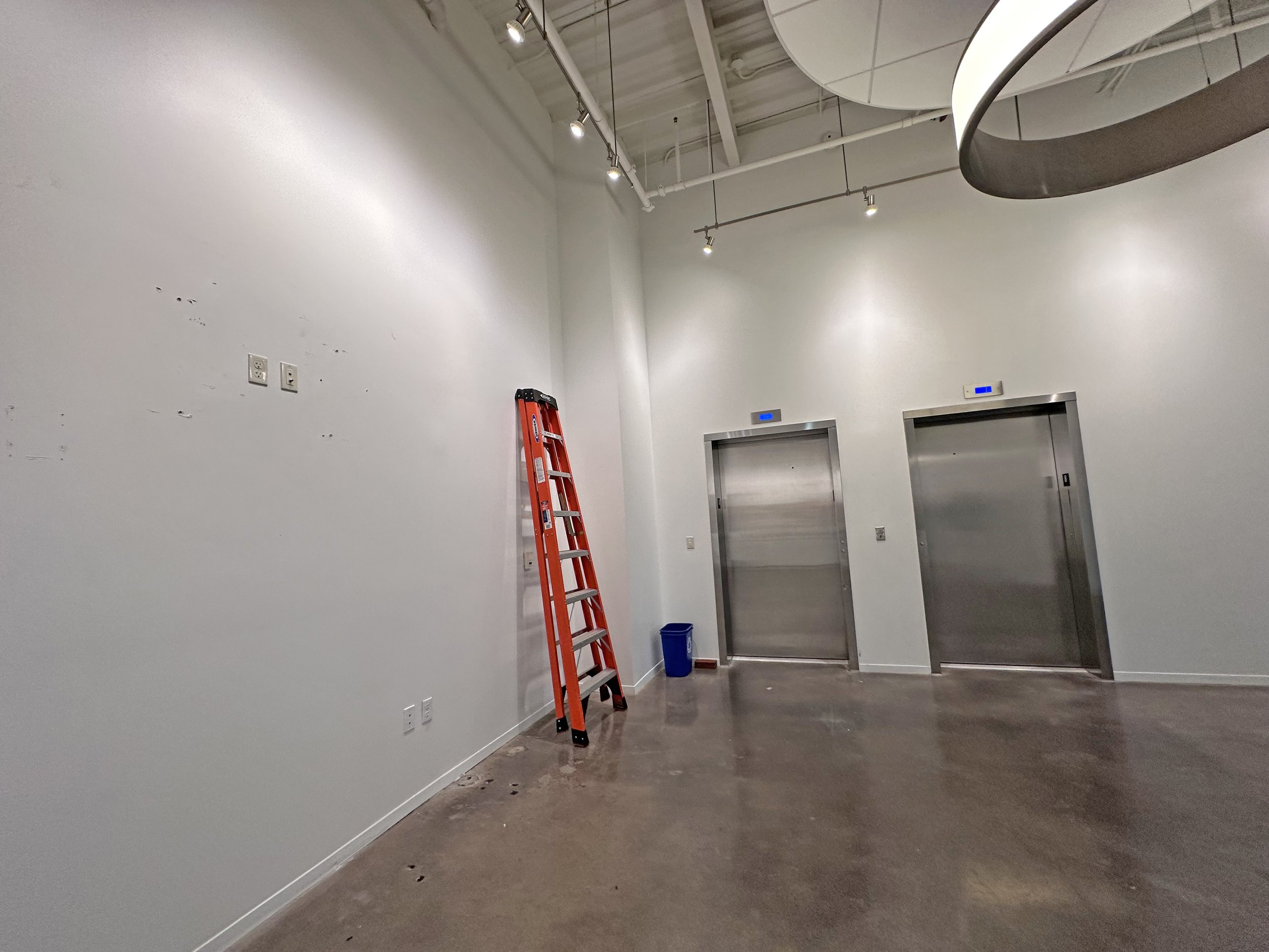 Empty room with white walls, polished concrete floor, two metal elevator doors with blue indicator lights, a red ladder leaning against the wall, a small blue trash bin, and visible ceiling track lighting.