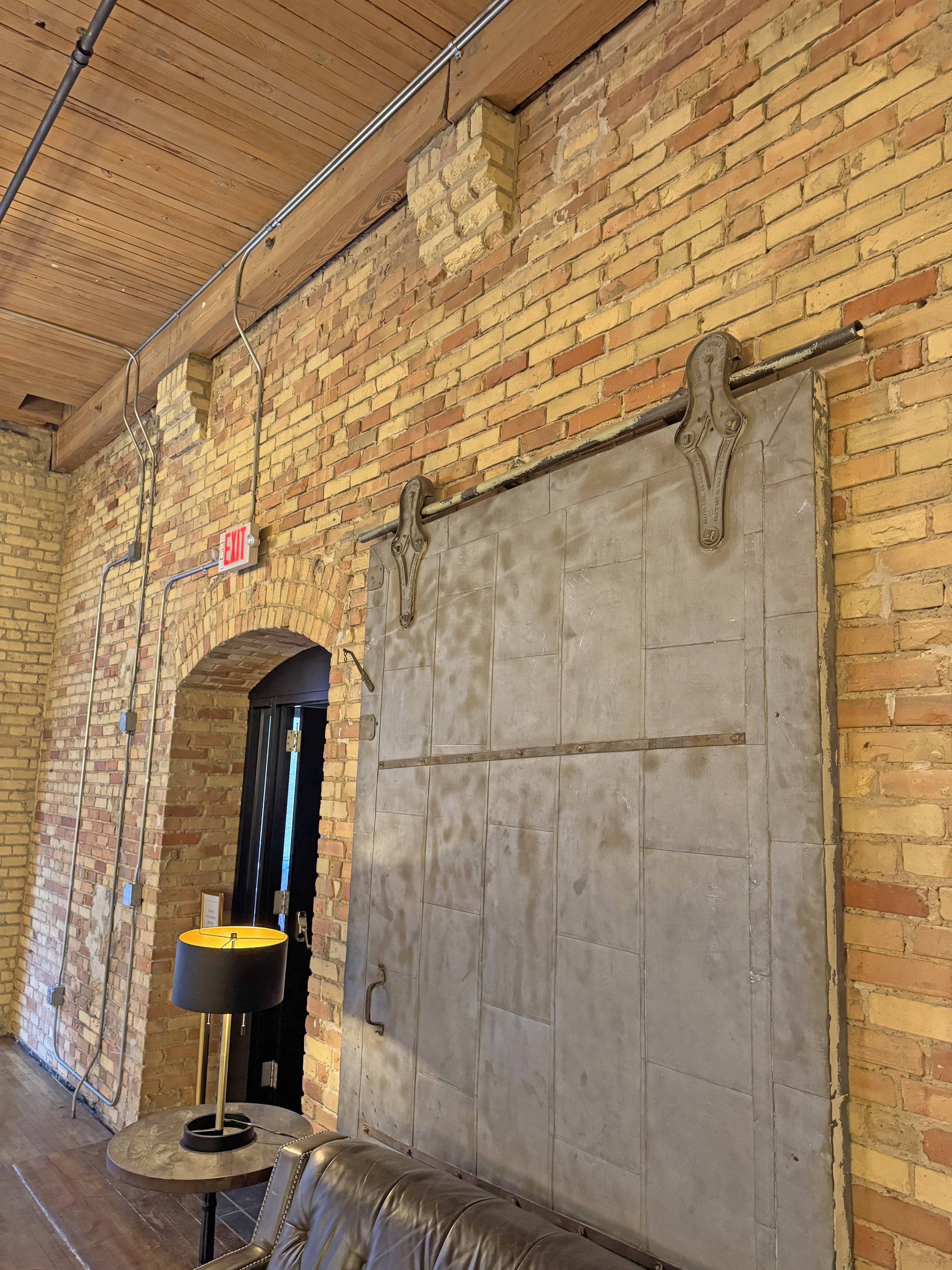 Interior with exposed brick walls, a sliding metal door on a track, and rustic decor including a table lamp and leather chair.