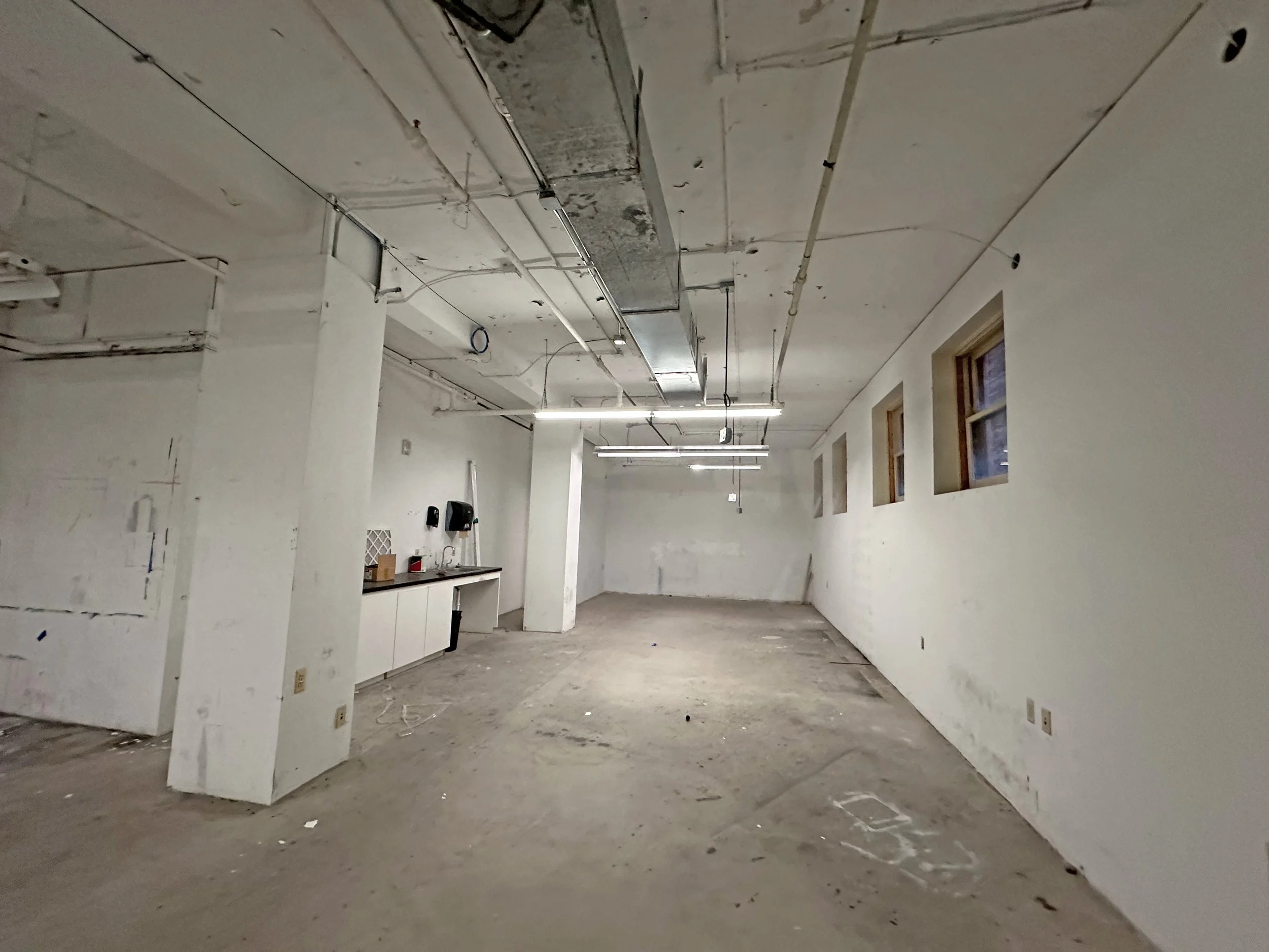 Empty unfinished room with white walls, exposed ceiling pipes, and a sink area on the left. There are four small windows on the right wall, and the floor is bare concrete with some markings and dirt.