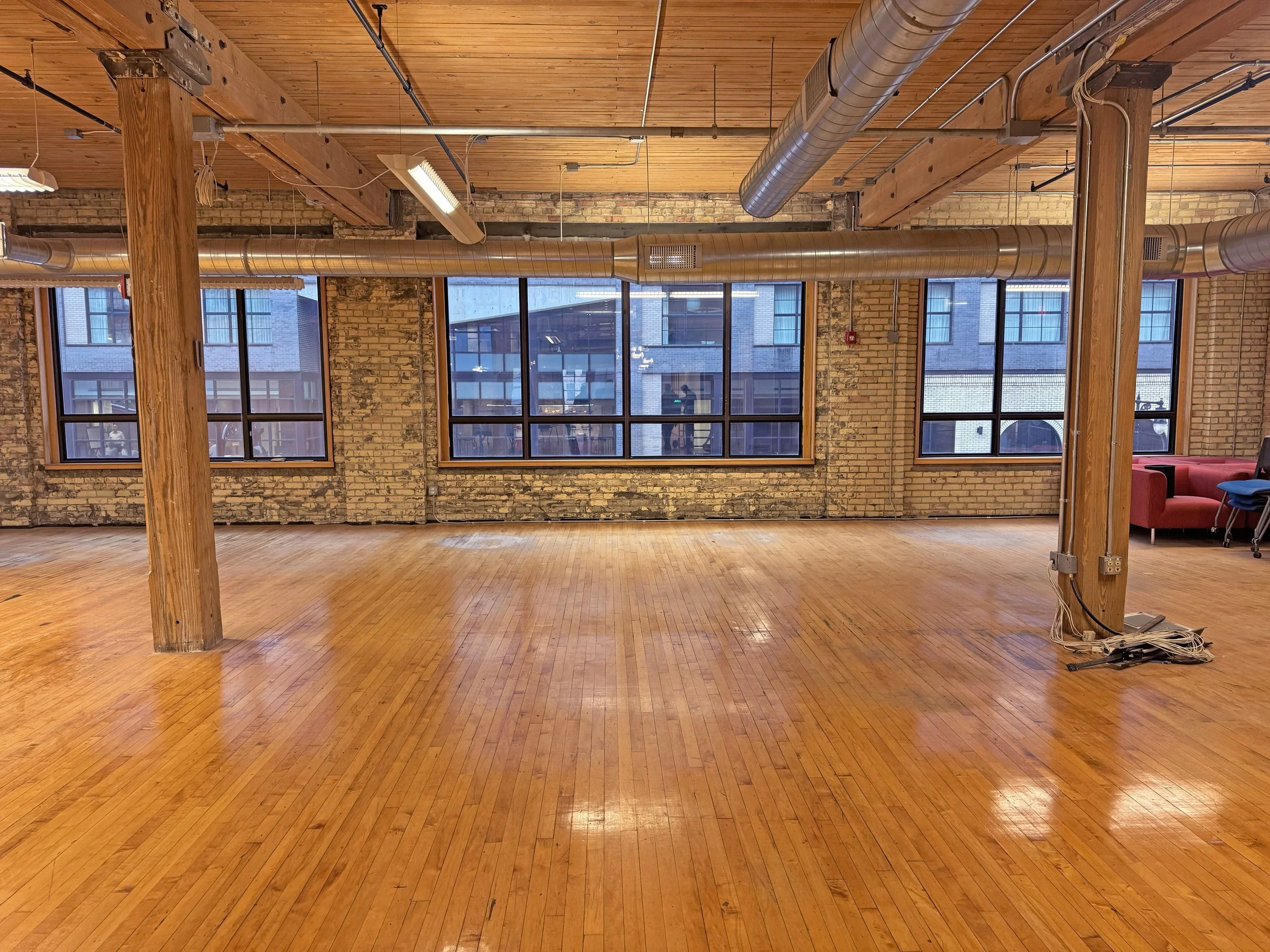 Empty loft-style room with wooden plank flooring, large windows, brick walls, and exposed air ducts. Chairs and a red sofa are in the corner.