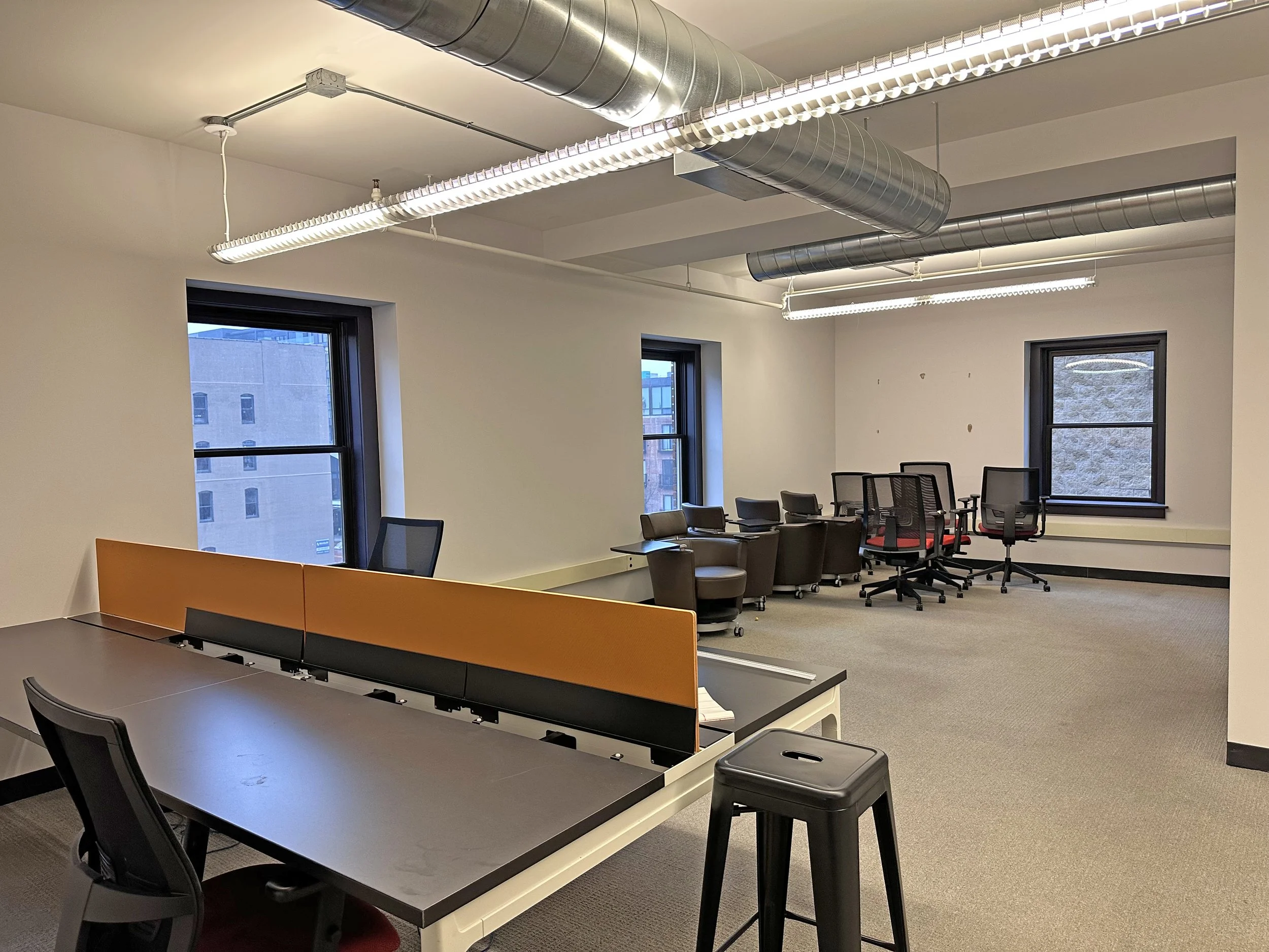 An office space with multiple workstations, chairs, and windows, featuring modern ceiling air ducts and lighting.