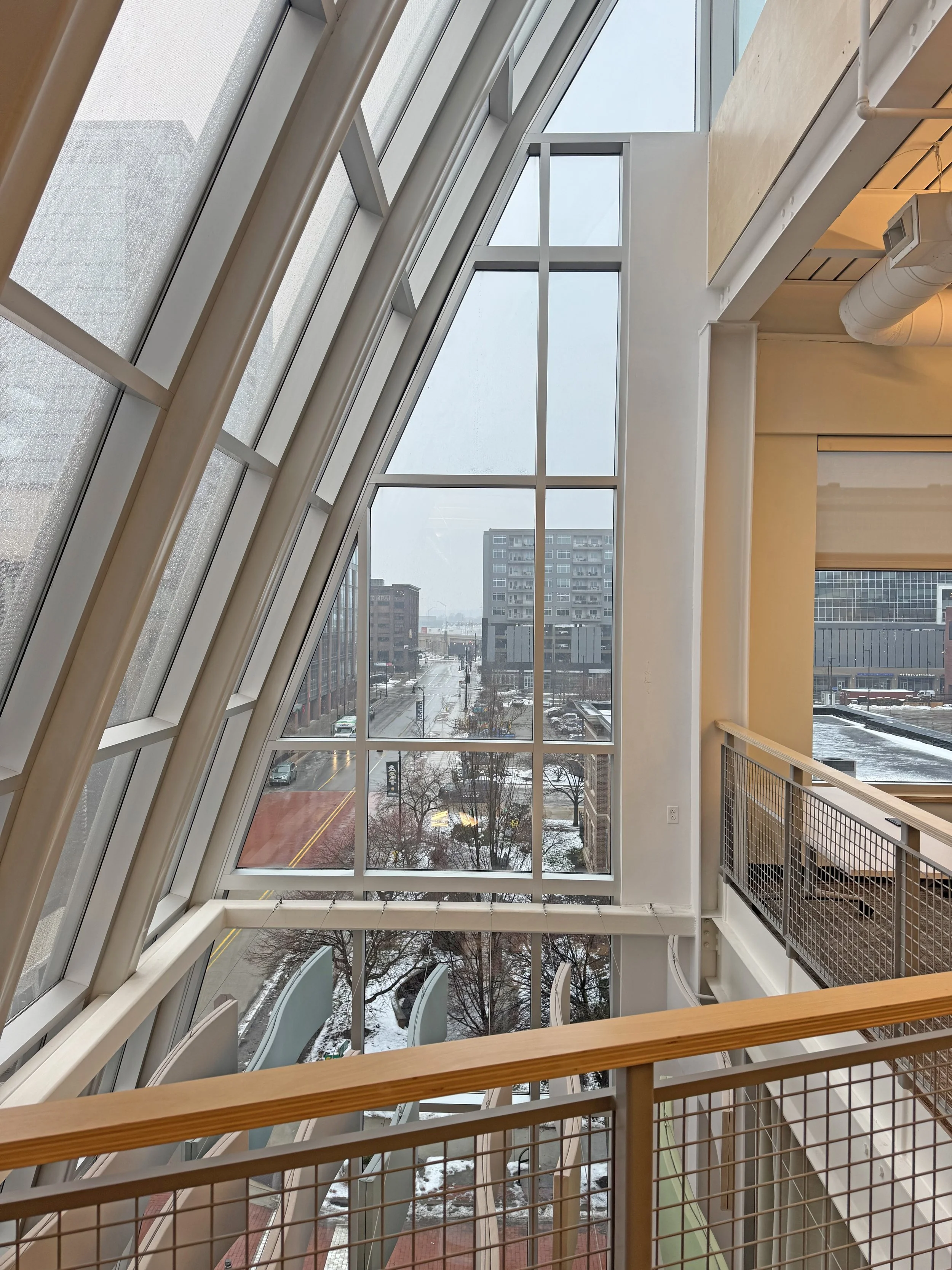 Interior of a modern building with large angled glass windows showing an urban street scene with cars, snow, and buildings outside.