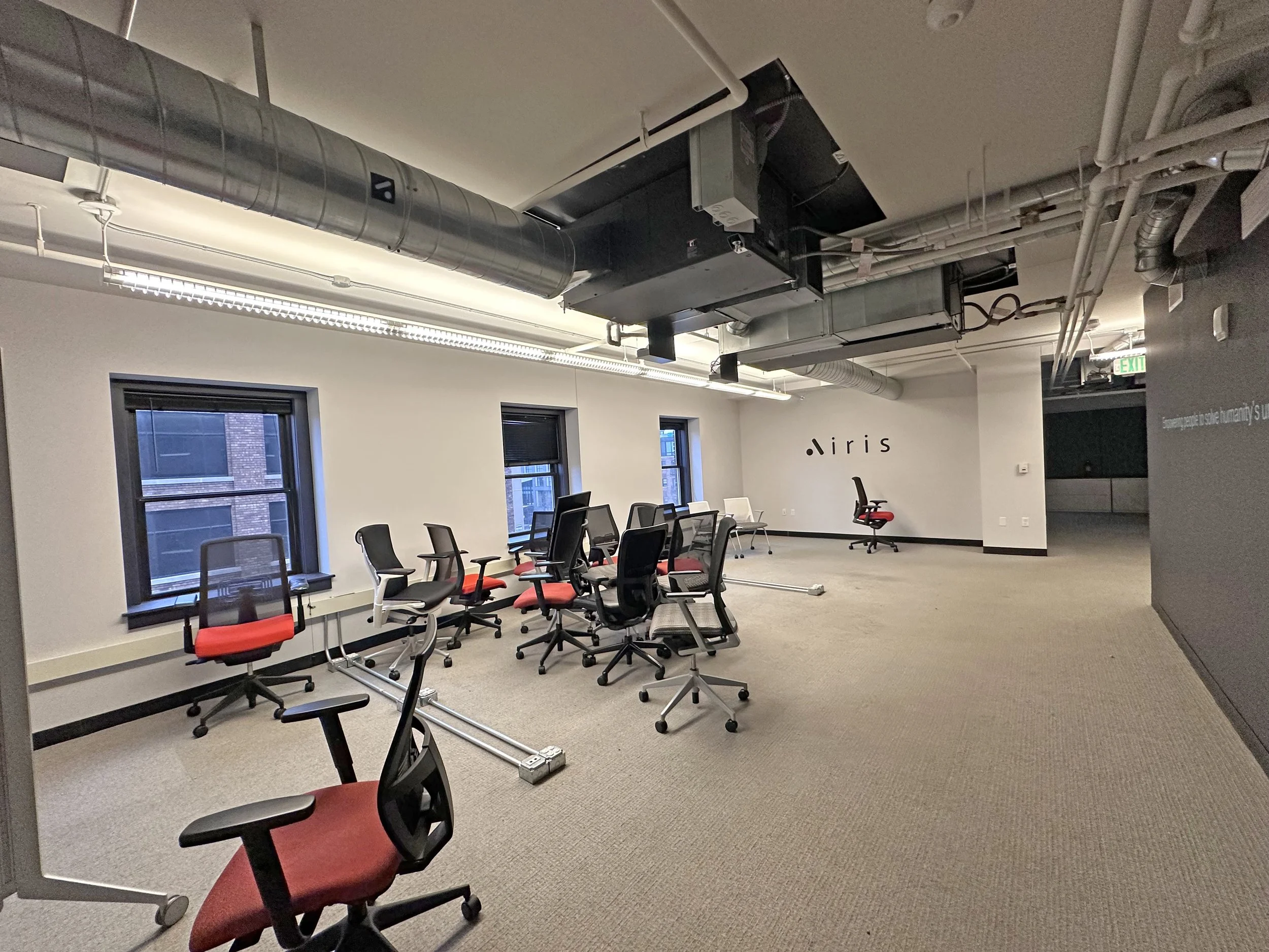Office space with multiple black and white office chairs with red cushions, some on rollers, arranged around a white table, near three windows. There are exposed ducts and pipes on the ceiling. The room has beige carpet, and a wall with the logo 'Airis' in black. An exit sign is visible in the corner.