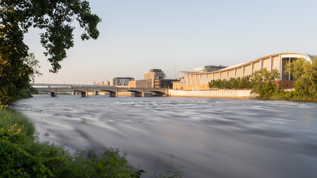 Cityscape with a river in the foreground and modern buildings in the background, with a bridge over the river and greenery on the sides.