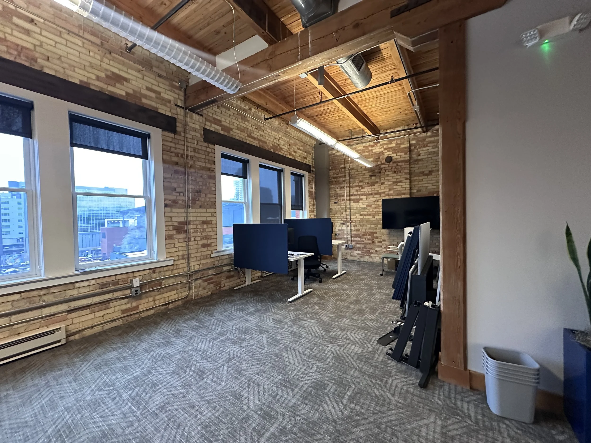 An office with exposed brick walls, multiple windows with blinds, and a wooden ceiling with visible beams and industrial-style ductwork. There are desks with blue privacy screens and a wall-mounted television or monitor. The floor is carpeted with a patterned design, and there are some folded display boards leaning against the wall.