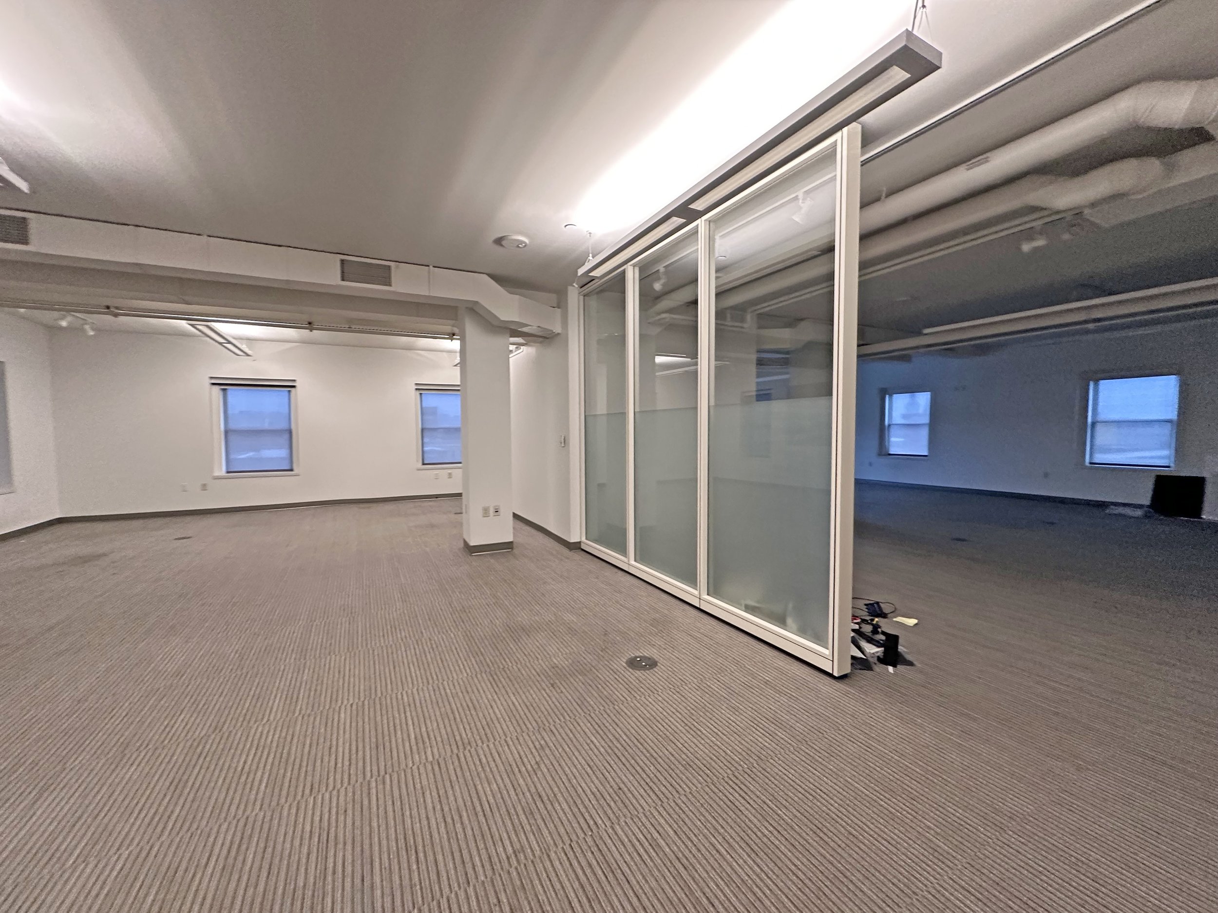 Empty interior office space with beige carpet, white walls, windows, and a glass partition.