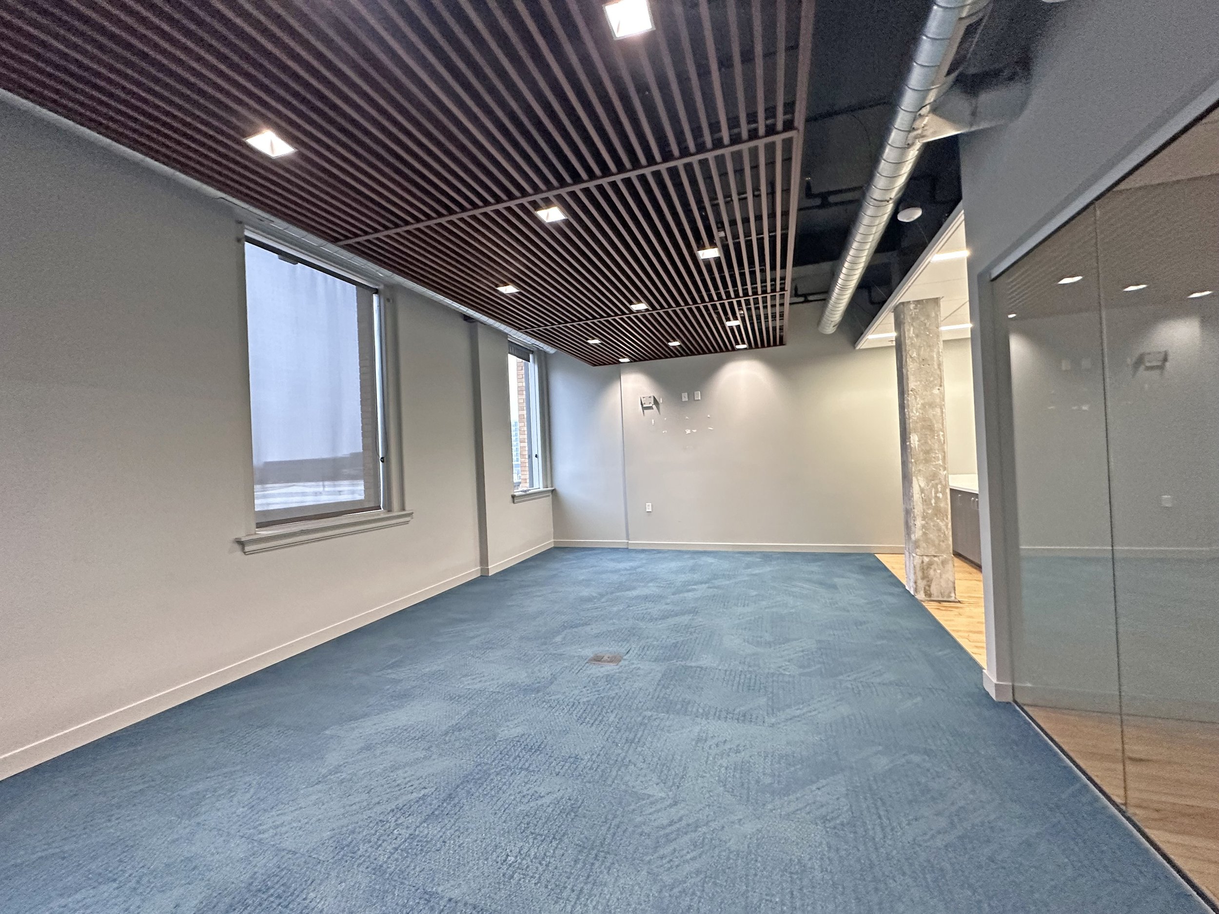 Empty office space with blue carpet, white walls, large windows, wooden ceiling, and a portion of glass wall or partition.