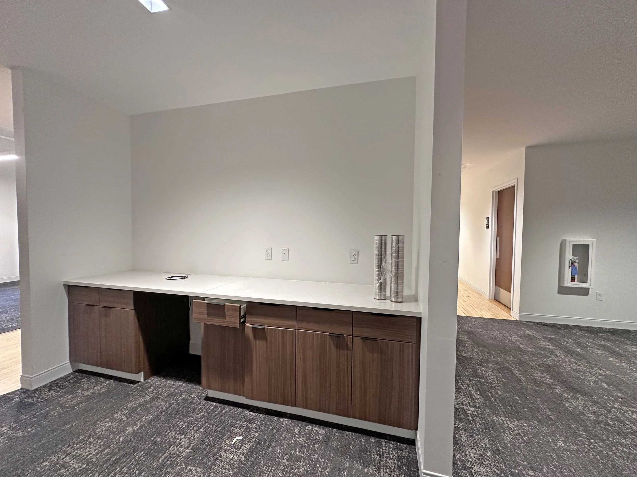 Office space with a long white countertop, wooden cabinets underneath, and three rolled-up silver posters on the side. There are electrical outlets on the wall, and a network cable on the counter.