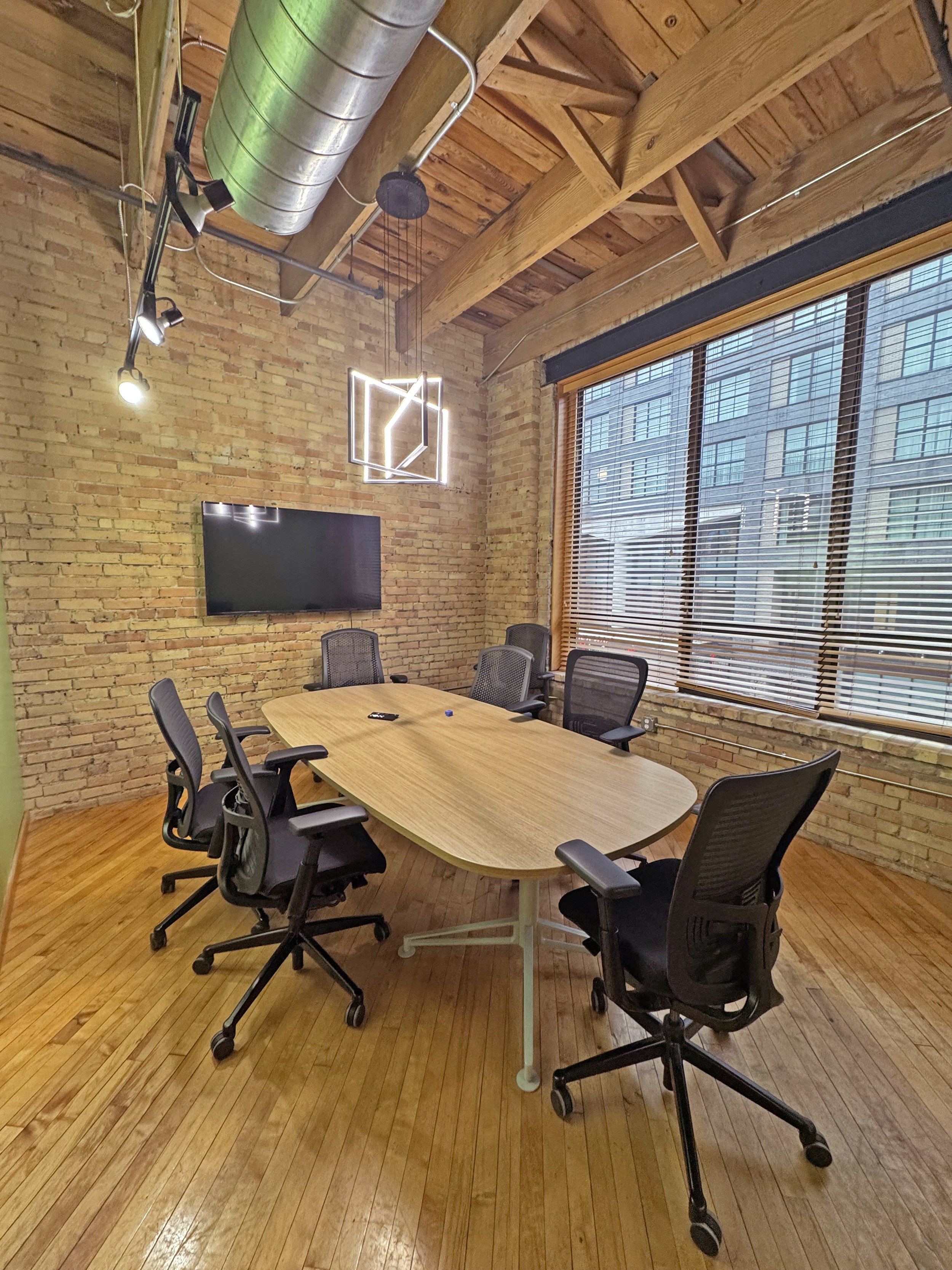 An empty conference room with a large wooden table, six black office chairs, a wall-mounted television, a brick wall, wooden ceiling beams, large windows with blinds, and a modern geometric light fixture.