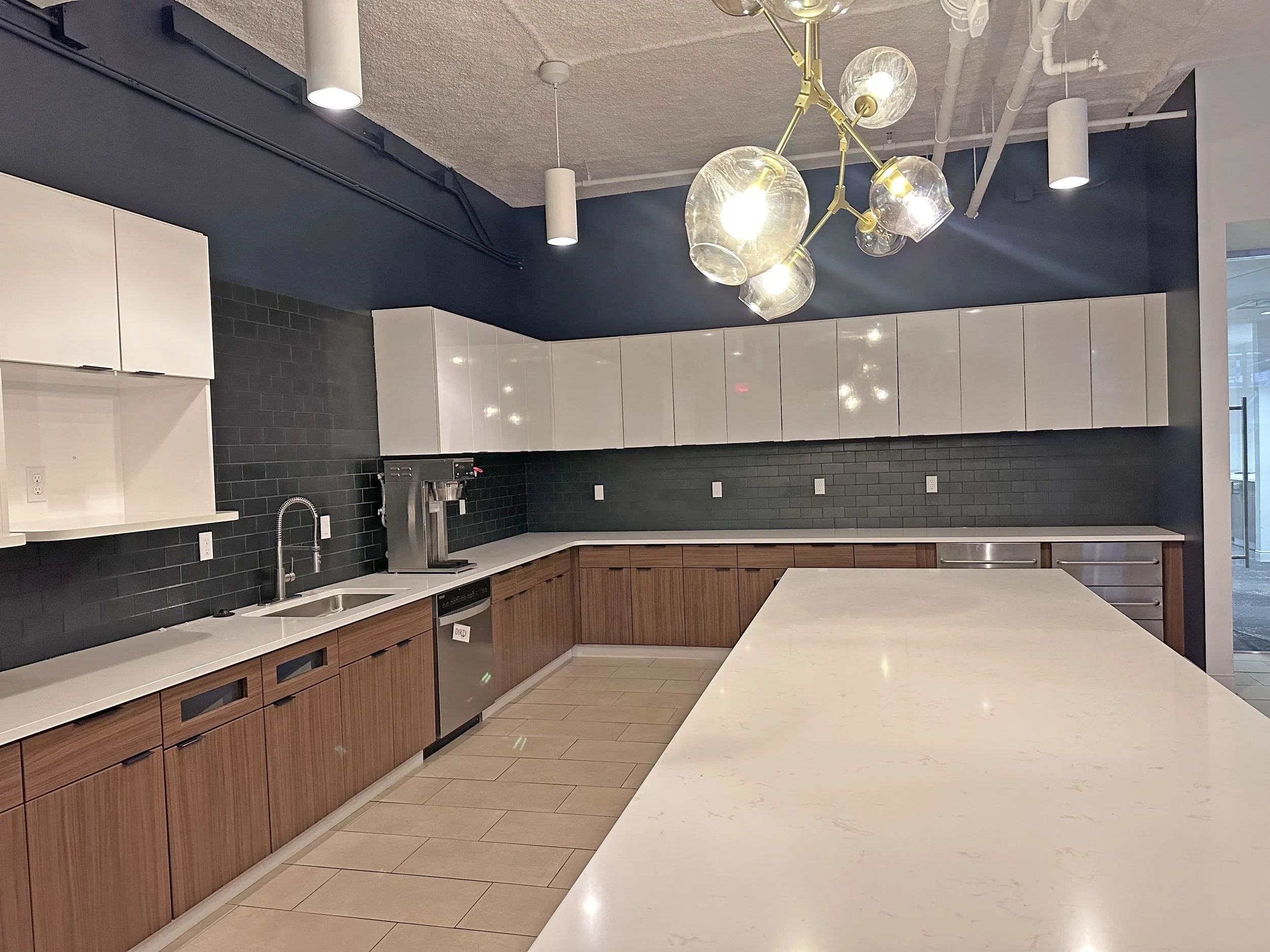 Modern kitchen with white countertops, wooden lower cabinets, white upper cabinets, a black backsplash, and a large central island. There is a chandelier with glass globes hanging from the ceiling.